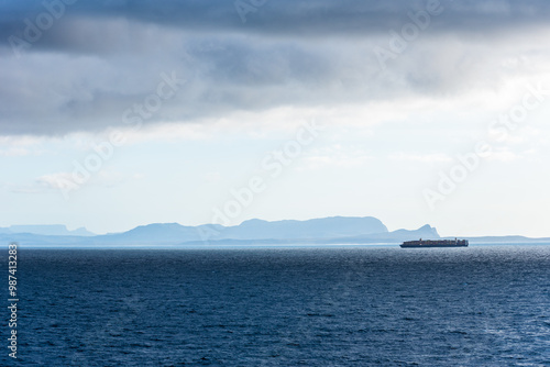 A misty horizon frames the rugged coastline near Cape of Good Hope, with a large container ship gliding along the shore. Sunlight breaks through clouds, casting a soft glow on the still dark waters.