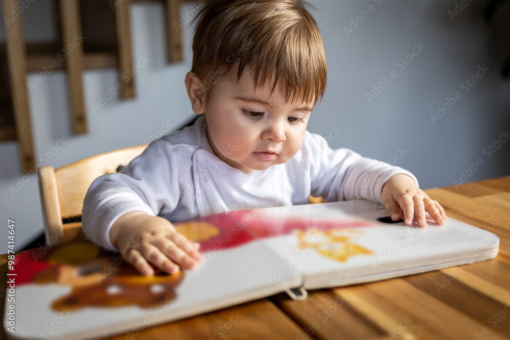 Little boy is looking through a children's book with stiff pages while sitting on a chair at the table Little boy is looking through a children's book with stiff pages while sitting on a chair at the table