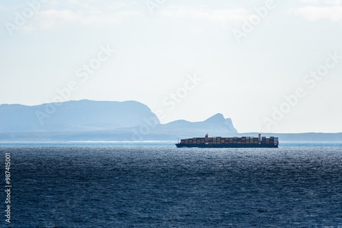 A misty horizon frames the rugged coastline near Cape of Good Hope, with a large container ship gliding along the shore. Sunlight breaks through clouds, casting a soft glow on the still dark waters.