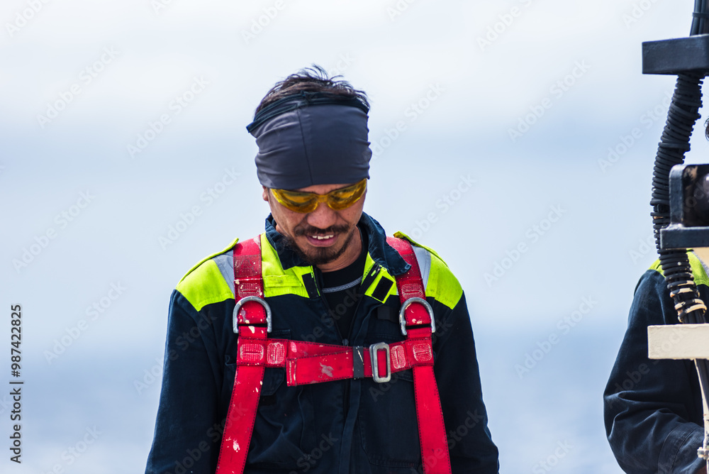 Standing on the deck of a massive container ship, a focused sailor secures his safety harness, preparing for the task ahead. Standing on the deck of a massive container ship, a focused sailor secures his safety harness, preparing for the task ahead.