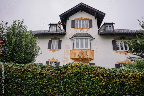 Bavaria, Germany - September 14, 2024 - An ornate, Bavarian-style house with painted details, a yellow balcony, and a hedge in the foreground on a cloudy day..