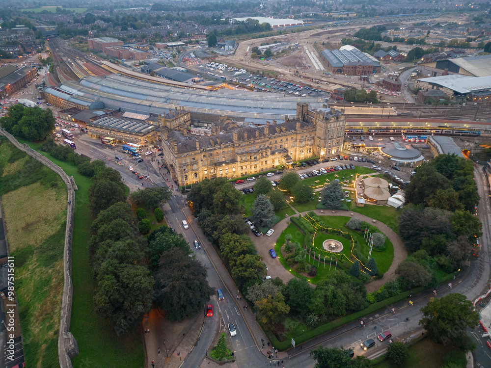 Beautiful illuminated York Railway Station and Principal York Hotel exterior at dusk. Aerial view of historic buildings and old city wall. Green park with trees and flag poles. Road with traffic.