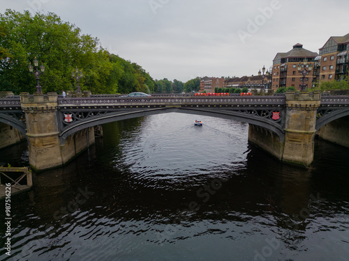 Closeup of historic Skeldergate Bridge in York, UK. Boat sailing under the bridge. Aerial view above river Ouse towards residential buildings and public park. Reflection of stone and metal bridge arch