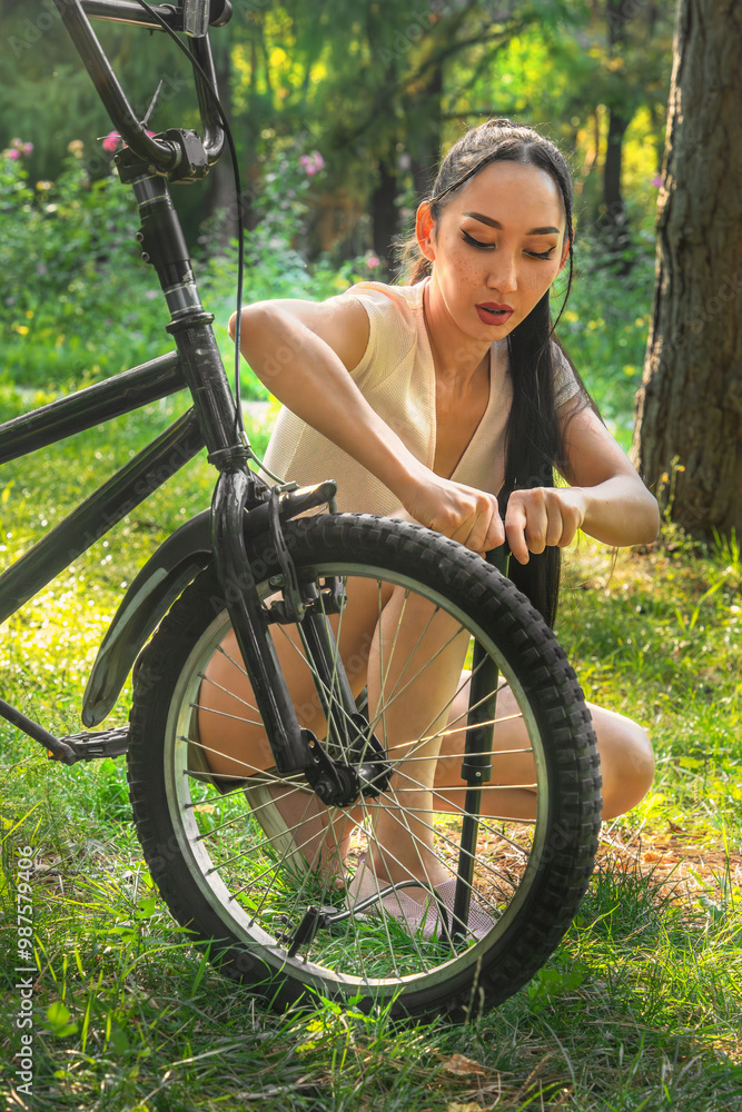 Fototapeta premium Beautiful Asian girl pumps a bicycle wheel with a pump in a park or forest at sunset. Bicycle repair. Healthy lifestyle.. Close-up