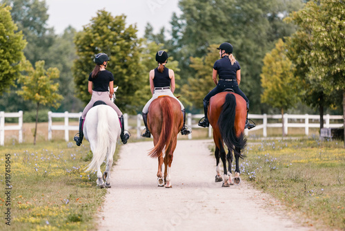 Rear view of three female riders riding horses side by side near white wood fencing, returning to the horse farm