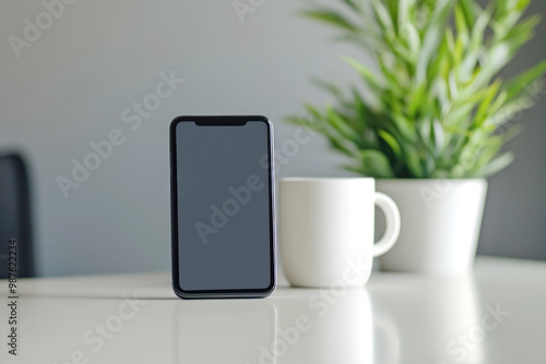 Black smartphone standing upright on a white table with a white mug and green plant in the background, creating a minimalist modern tech scene.