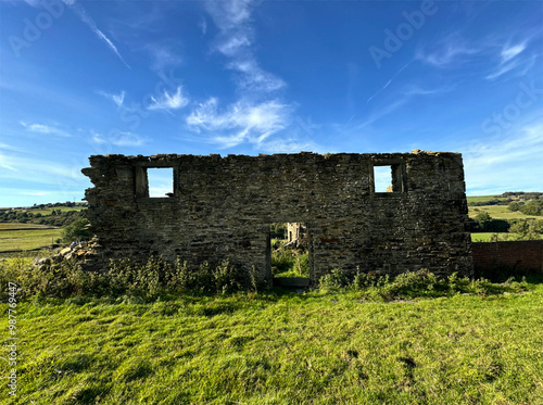 A dilapidated stone farmhouse rises from a grassy field, set against a clear blue sky. The nearby landscape boasts undulating hills and verdant greenery close to Goose Eye, Keighley, UK. photo