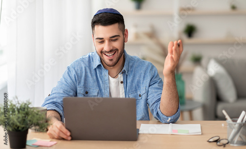 Distant Communication Concept. Portrait of happy jewish male in yarmulke and casual shirt making video call sitting at desk at home office and talking with colleagues, students or family, explaining photo