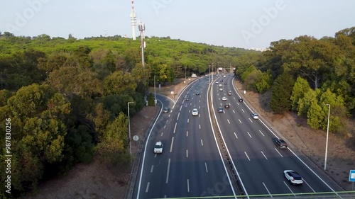 Autoestrada em Monsanto, Lisboa, ao pôr do sol com tráfego em movimento. O céu alaranjado ilumina a estrada e os veículos, criando um contraste entre a natureza e a paisagem urbana.