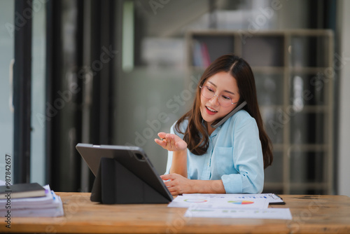 Businesswoman on Phone Call: A focused businesswoman, dressed professionally in a blue blouse, multitasks on a phone call while reviewing documents on her tablet at her desk. The soft lighting of the 