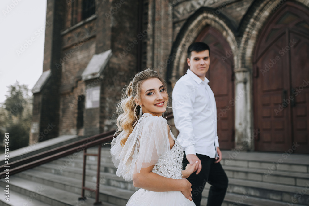 Fototapeta premium Fashionable young groom and beautiful smiling bride curly blonde in white lace dress hug, walk together against the background of old retro church. Wedding photo, portrait of happy newlyweds.