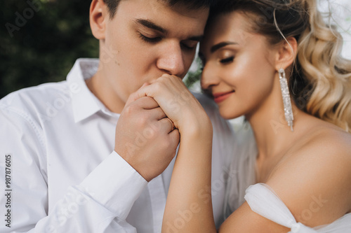 Young stylish groom tenderly kisses hand of smiling beautiful blonde bride in white dress. Close-up wedding photo, portrait of happy newlyweds.