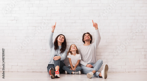 Fényképezés Family of three sitting on floor near white brick wall, pointing and looking upw