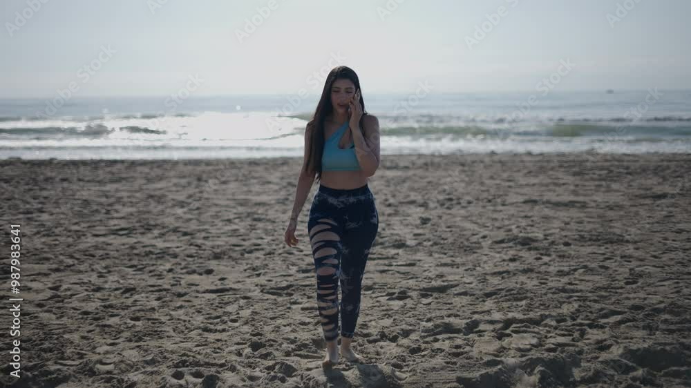 Young girl with long hair brunette Caucasian woman walking facing camera on the sand of the beach on a sunny day makes a call on her smartphone Young girl with long hair brunette Caucasian woman walking facing camera on the sand of the beach on a sunny day makes a call on her smartphone
