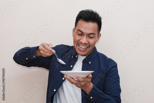 A Southeast Asian man happy wearing a blue shirt while holding an empty plate and spoon with happy and excited expression. A man showing and pointing empty mockup plate. photo