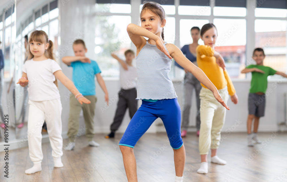 Fototapeta premium Positive juvenile girl engaged in hip-hop dance together with children's group and female trainer in training room