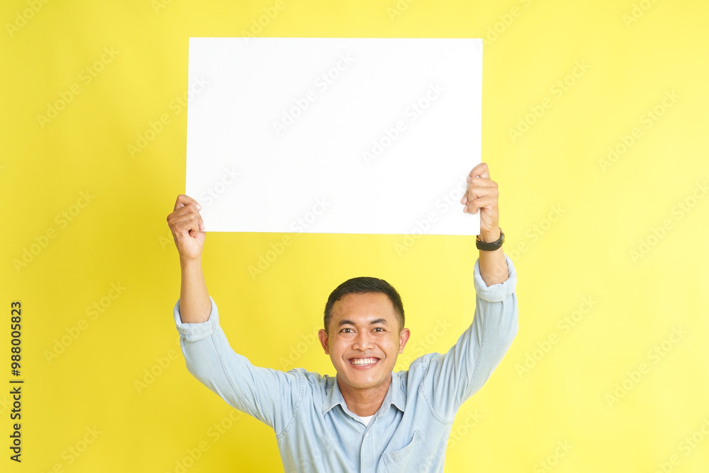 Asian man in light blue denim shirt holding blank white board for advertisement isolated on yellow background Asian man in light blue denim shirt holding blank white board for advertisement isolated on yellow background