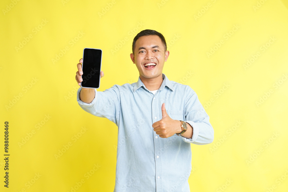 Asian man in light blue denim shirt holding blank screen phone and recommended gesture isolated on yellow background Asian man in light blue denim shirt holding blank screen phone and recommended gesture isolated on yellow background