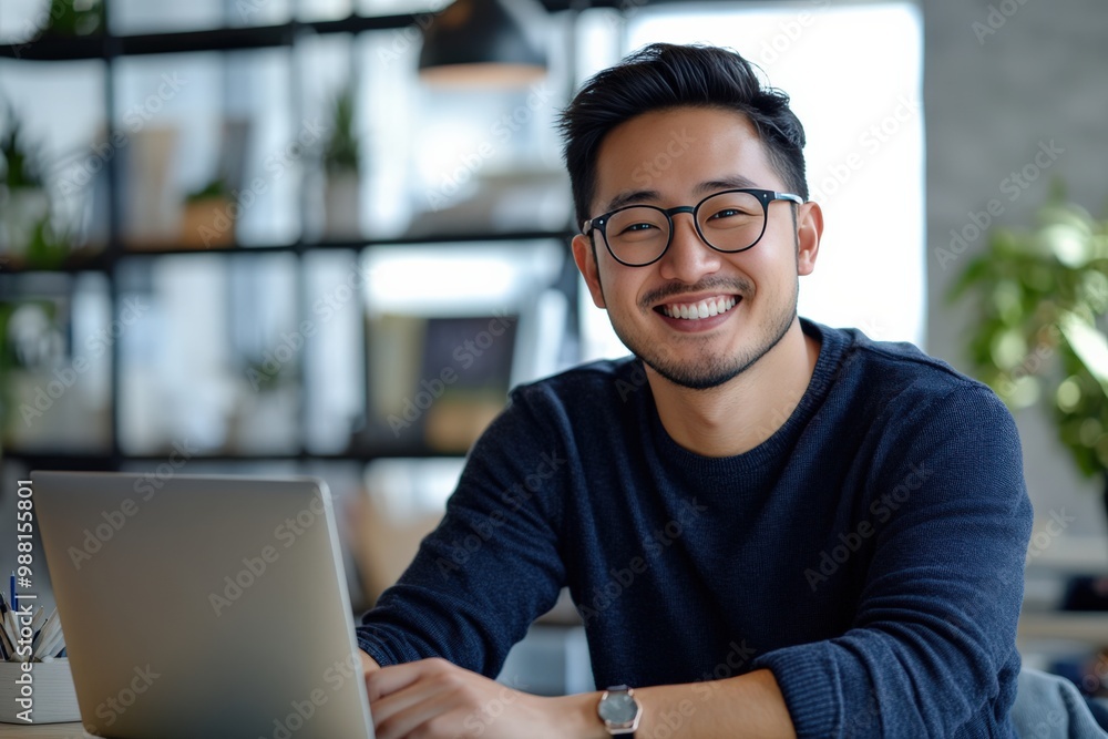 Smiling Successful Asian Man Sitting at Her Desk Working on a Laptop Computer Smiling Successful Asian Man Sitting at Her Desk Working on a Laptop Computer