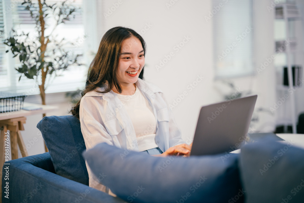 Young asian woman happily works from home, smiling and typing on her laptop on a cozy blue couch. She balances work and personal life seamlessly in a modern setting Young asian woman happily works from home, smiling and typing on her laptop on a cozy blue couch. She balances work and personal life seamlessly in a modern setting