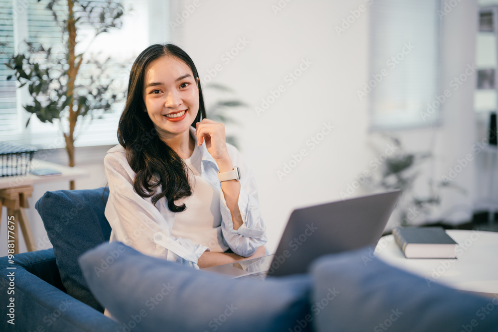 Young asian freelancer is working from her home office, sitting on a comfortable blue couch and using her laptop. She is happy and smiling