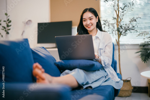 Happy asian woman working from home on her laptop, sitting comfortably on a sofa, embodying a modern lifestyle