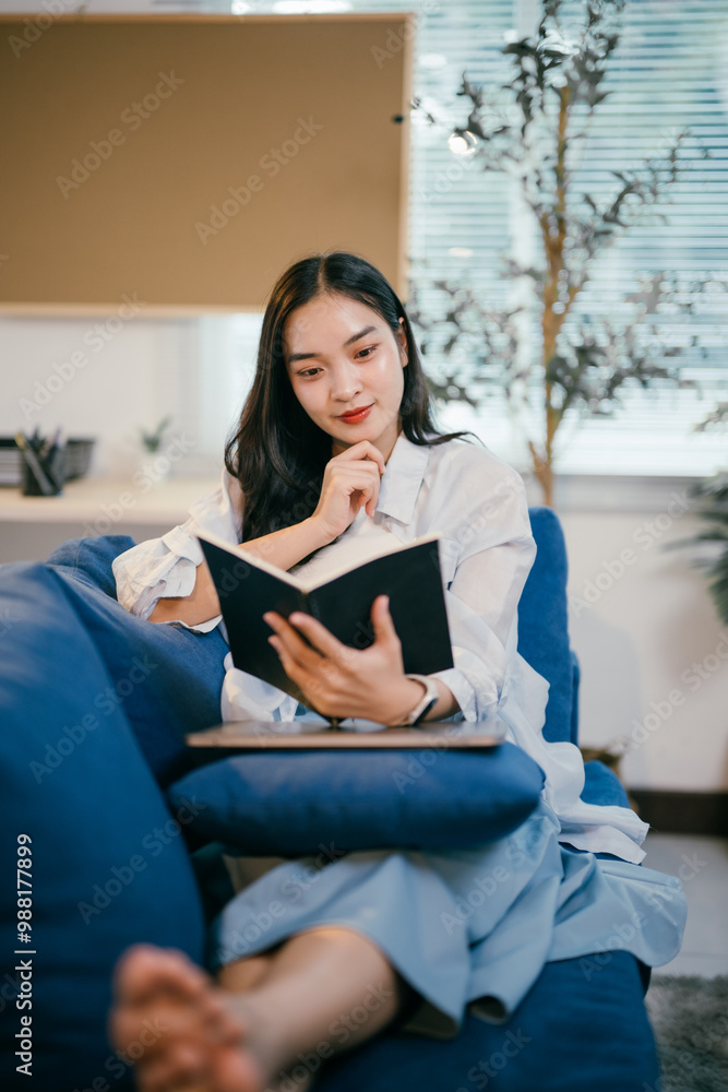 Young woman is sitting on a comfortable blue sofa in her living room, engrossed in a captivating book, taking a break from her busy schedule Young woman is sitting on a comfortable blue sofa in her living room, engrossed in a captivating book, taking a break from her busy schedule