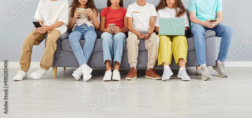 Cropped photo of a group of young school children in casual clothes sitting on sofa in a row using laptop with mobile phones isolated on gray background. People and technology concept.