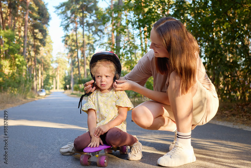 Mother teaching her toddler how to ride penny board on the asphalt in park, carefully guiding the little kid enjoying an active summer day together outdoors