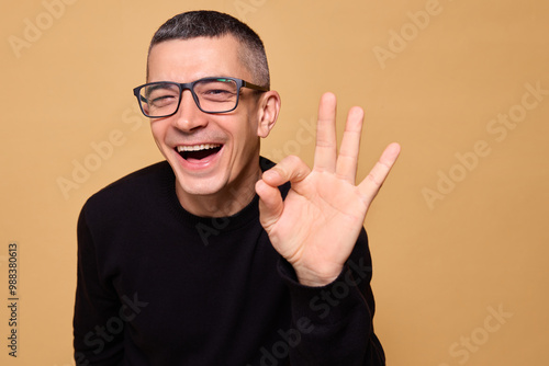 Cheerful funny man wearing black raglan and glasses showing ok sign with big smile expressing approval and happiness giving playful gesture with fingers isolated over beige background