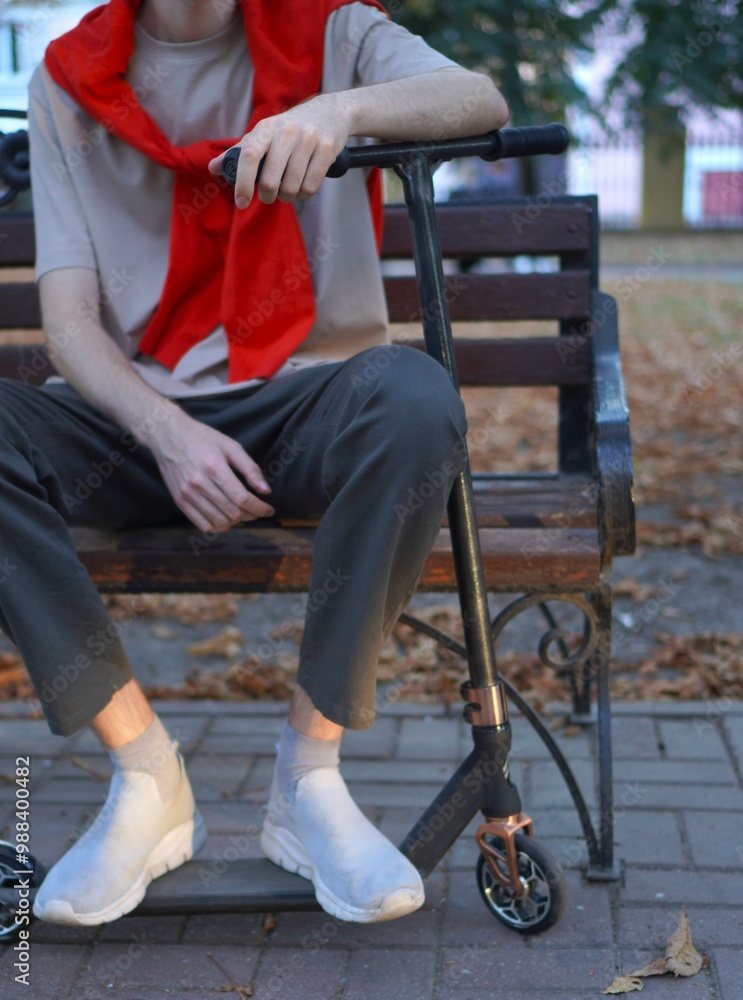 Obraz premium Golden autumn with a young athlete in a red hoodie on his shoulders, sitting on a bench with a scooter in a city park.