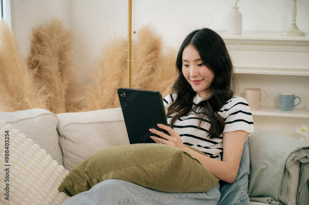 A beautiful woman sits on a large, comfortable sofa by the window of her house. She plays social media on tablet during the weekend. A beautiful woman sits on a large, comfortable sofa by the window of her house. She plays social media on tablet during the weekend.