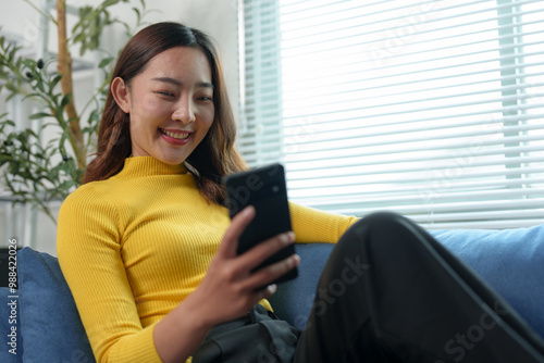 Happy attractive Asian woman holding mobile phone using technology for online shopping, e-commerce, buying things online, texting, greeting, chatting, relaxing on sofa in living room at home.
