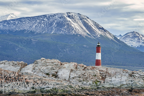 Les Eclaireurs Lighthouse :  Red and white lighthouse on rocky outcrop, snow-capped mountains in background, partly cloudy sky, Ushuaia’s maritime charm and natural beauty photo