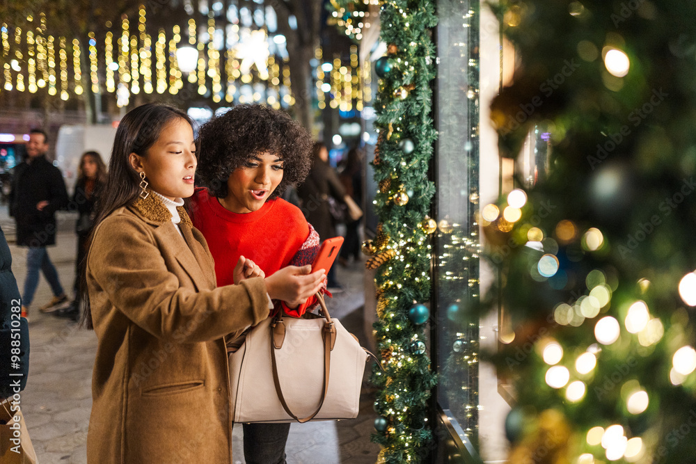 Obraz premium Two young women window shopping while checking smartphone in a decorated city at christmas time
