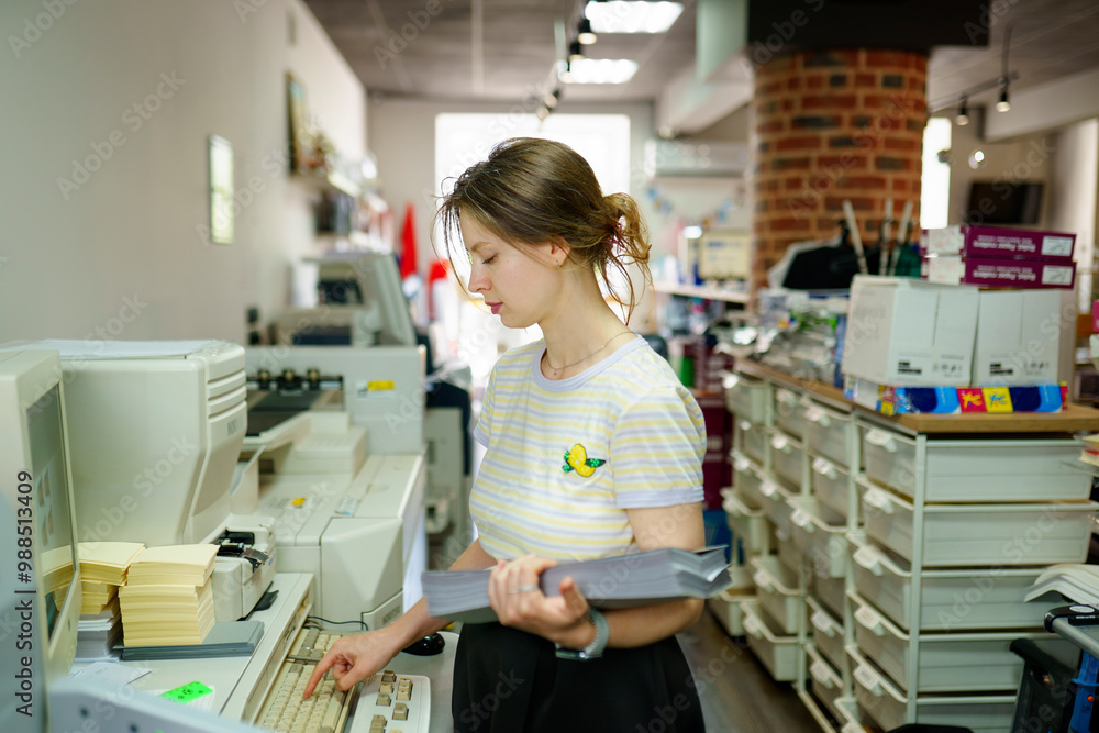 Woman holding stack of papers and using computer in a printing shop Woman holding stack of papers and using computer in a printing shop