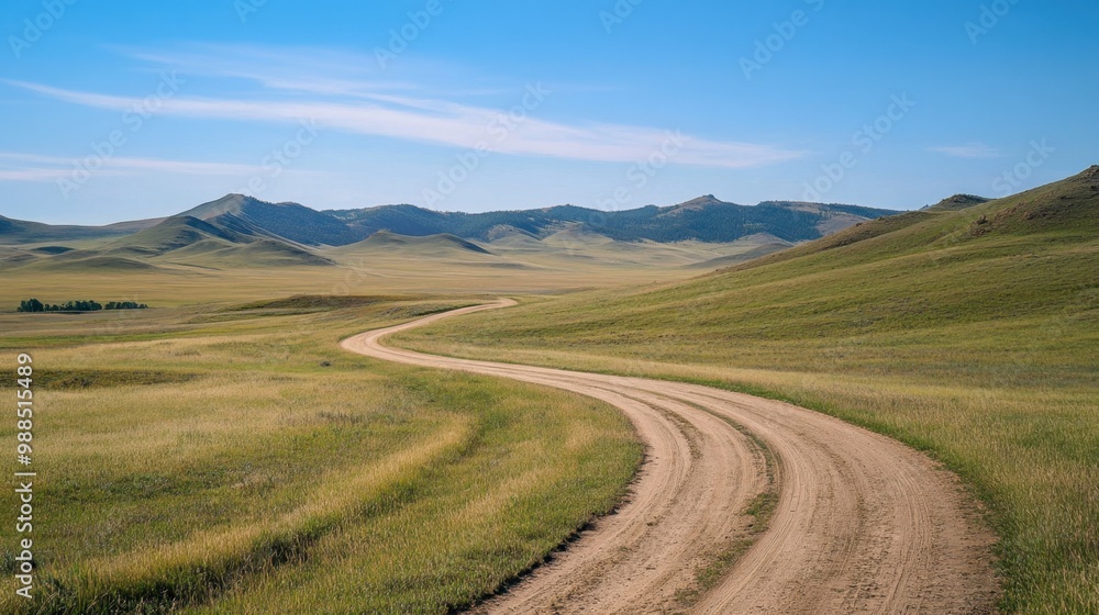 Fototapeta premium A winding dirt road stretches across open grasslands with mountains in the background and a clear blue sky overhead