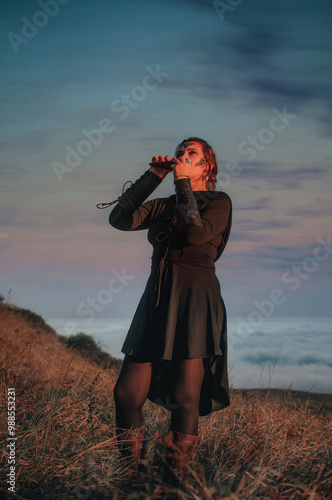 Beautiful girl in gothic style clothes with tattoo on her face plays ocarina in the mountains