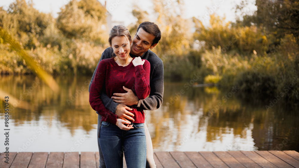 Fototapeta premium Smiling man hugging woman on pier near pond in autumn
