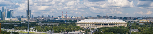 The iconic Luzhniki Stadium is nestled within Luzhniki Park, with the modern Moscow City skyline rising in the background under blue skies