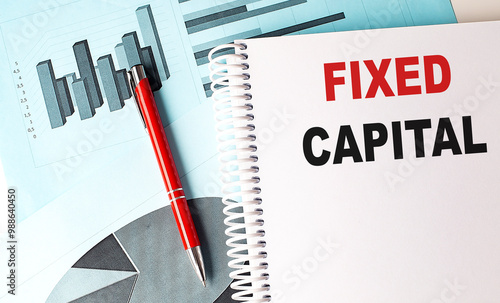 Notebook with the text fixed capital on a white cover, placed on financial charts with a red pen