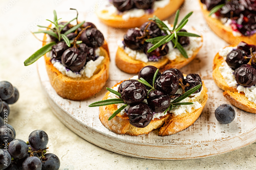 White board with Roasted Grape and Goat ricotta Cheese Crostini with rosemary. Fresh grape plant on the side. White board with Roasted Grape and Goat ricotta Cheese Crostini with rosemary. Fresh grape plant on the side.