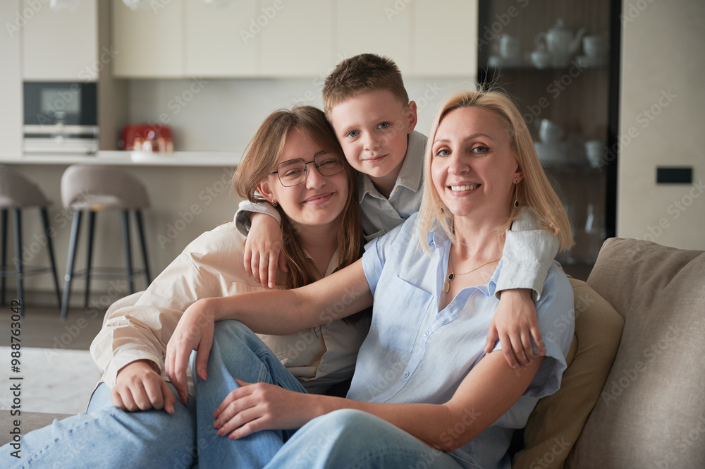 Lovely family relaxing on couch in living room. Little boy hugging his sister and mom. Cozy spending time with family indoors. In background, modern kitchen, neutral colors, minimalistic design. Lovely family relaxing on couch in living room. Little boy hugging his sister and mom. Cozy spending time with family indoors. In background, modern kitchen, neutral colors, minimalistic design.
