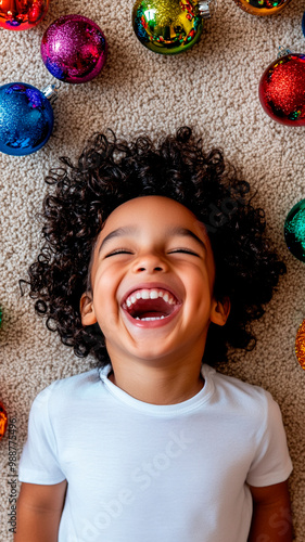 Un niño de pelo oscuro y rizado, vestido con una camisa blanca, ríe rodeado de coloridos adornos navideños sobre una alfombra beige. La escena está captada desde arriba a pantalla completa, mostrando  photo