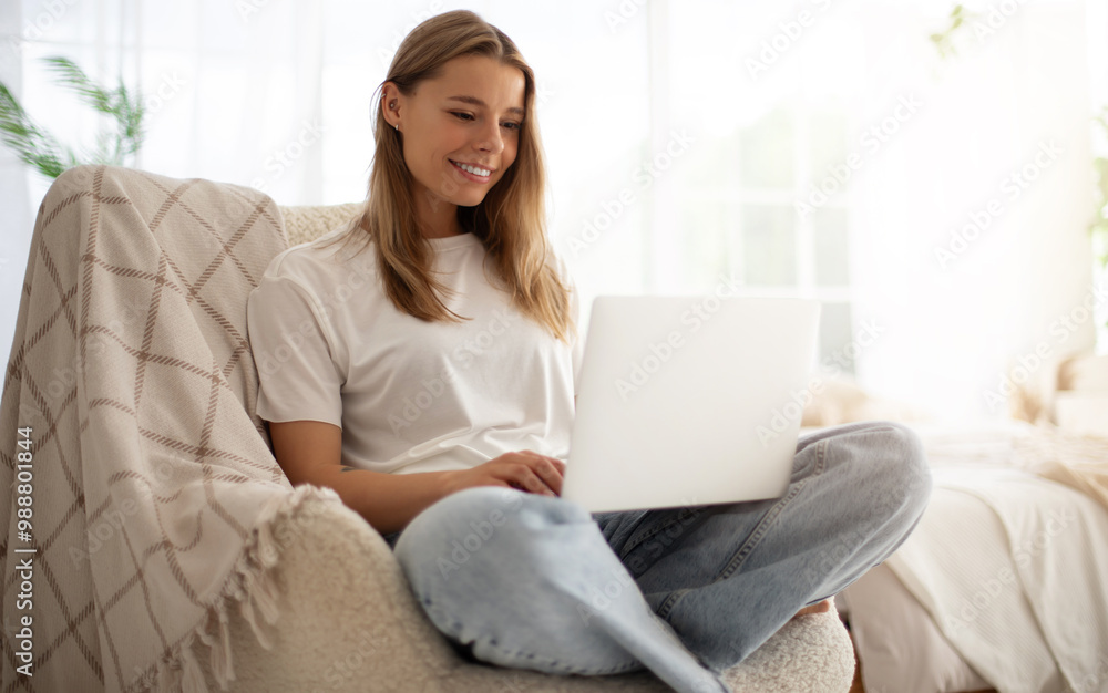 Young woman sits comfortably in cozy chair, working on her laptop in bright, modern home setting. She is smiling, dressed casually in jeans and t-shirt, surrounded by natural light and indoor plants Young woman sits comfortably in cozy chair, working on her laptop in bright, modern home setting. She is smiling, dressed casually in jeans and t-shirt, surrounded by natural light and indoor plants