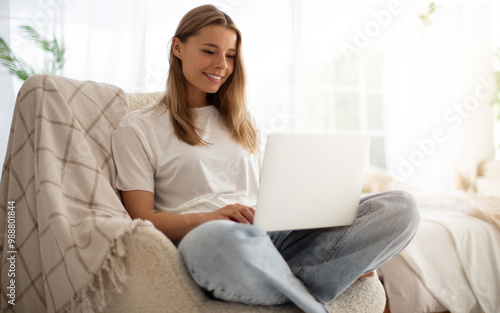 Young woman sits comfortably in cozy chair, working on her laptop in bright, modern home setting. She is smiling, dressed casually in jeans and t-shirt, surrounded by natural light and indoor plants