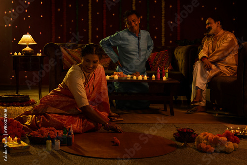 Woman decorating floor with flowers on the occasion of Diwali and two men watching and enjoying while sitting on sofa at home photo