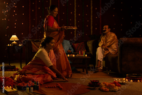 Two senior women decorating floor with flowers and diyas on the occasion of Diwali and their husband watching and enjoying while sitting beside on sofa at home photo