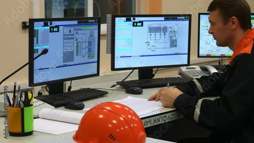Engineer is managing complex industrial system in control room while multitasking with paperwork and computer screens. Engineer in industrial control room is filling out paperwork while monitoring