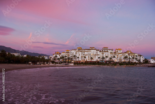 A beautiful coastal town is illuminated by a stunning pink and purple sunset. White buildings with yellow roofs stand out against the backdrop of serene seaside surroundings  in Puerto Banus Andausia photo